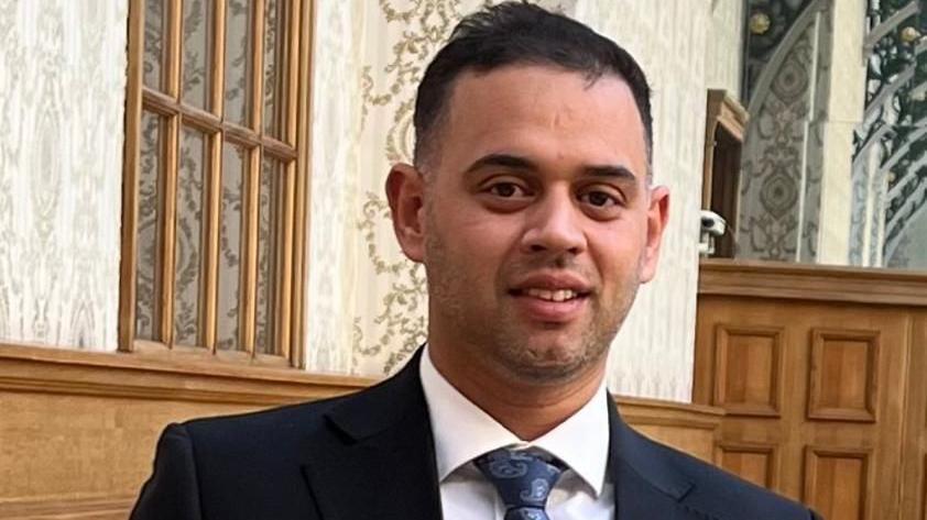 A close-up showing head and shoulders of a man with short black hair in a white shirt, navy jacket and patterned blue tie. He is standing inside a building with wooden wall-panels and cream and green patterned wallpaper.