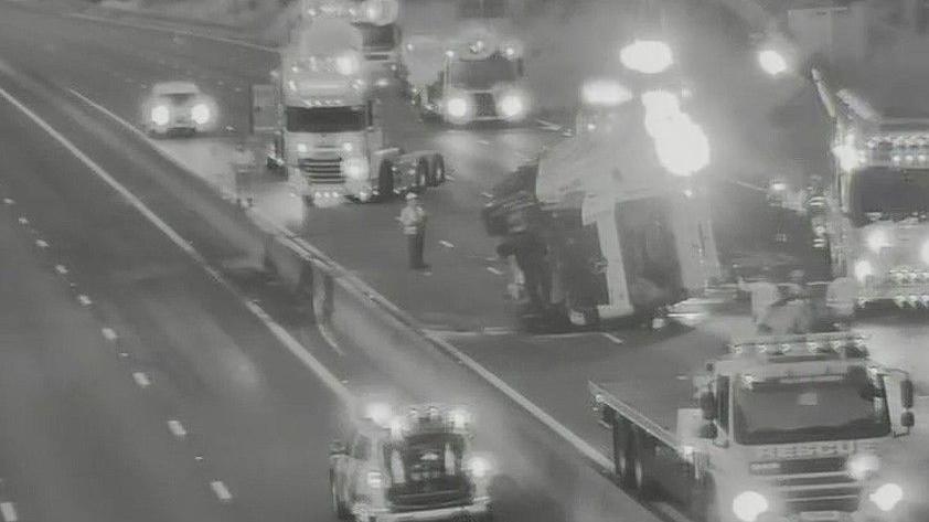 A screen capture of black and white CCTV showing a lorry overturned on a motorway carriageway. Emergency vehicles, including cars, ambulance and recovery trucks can be seen around it, with lights illuminated