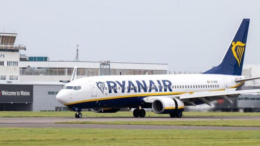 A Ryanair plane taxing at Cardiff Airport, with the terminal building in the background and a sign saying "Welcome to Wales" written at the left.