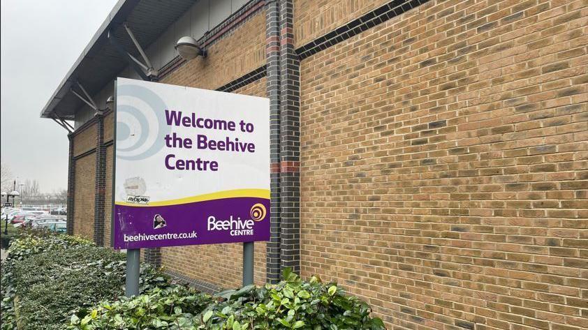 A white, yellow and purple sign next to a brick building, which reads: "Welcome to the Beehive Centre"