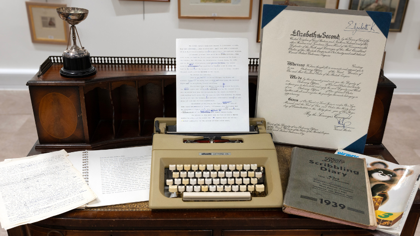 A wooden desk displaying items from the private collection, including a silver golfing trophy, a vintage typewriter, a scribbling diary, and typewritten notes.