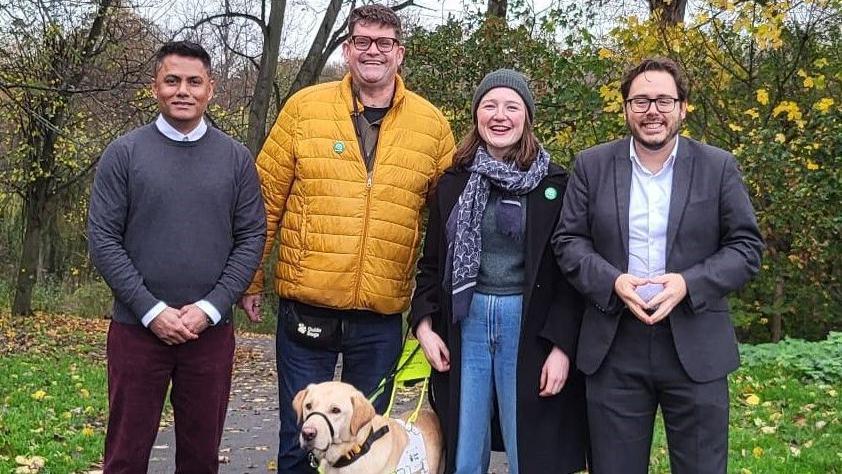 (Left to right) Khaled Musharraf, Alistair Chisholm, Sarah Peters, and Nick Hartley are standing in a leafy park and all smiling at the camera.
