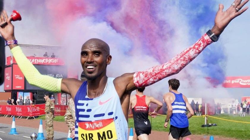 Sir Mo celebrates at an event with pink and blue smoke behind him. Two runners are also passing behind him. He is holding his arms up and is wearing a bib that says "Sir Mo" on it.