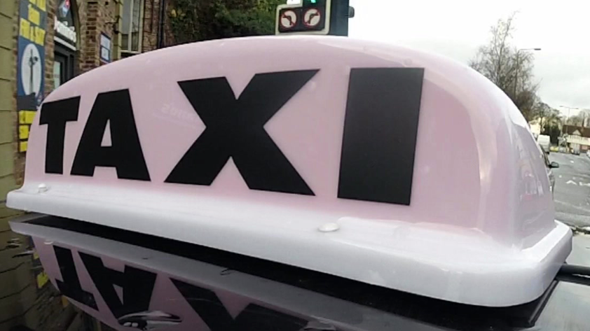 The picture shows the roof sign of a taxi. The sign is white with bold black letters spelling “TAXI.” It appears to be mounted on top of a vehicle and the background shows a street scene with traffic lights, cars, and buildings. The photo seems to have been taken during the day under cloudy weather conditions.