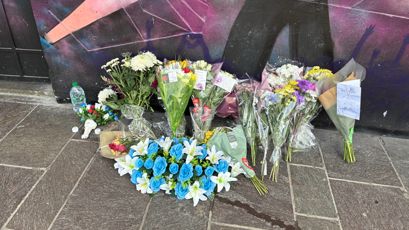 A number of bunches of flowers on a pavement next to a wall which has been painted with a bright, multi-coloured scene of people dancing.