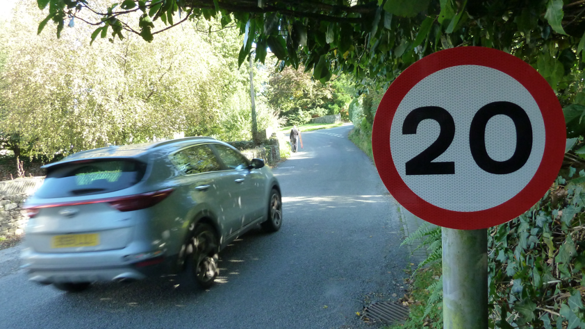 A grey car is driving past a 20mph circular speed restriction sign. A pedestrian is walking in the distance.