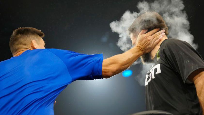 A photo from a slap fight in Las Vegas in May 24 between Wesley "All the Smoke" Drain and John "The Machine" Davis. The photo shows a man in a blue T-shirt slapping a bearded man in a black T-shirt. The slapper appears to hit his competitor with the full force of his flat hand. Chalk dust from the impact of the slap rings his head. 