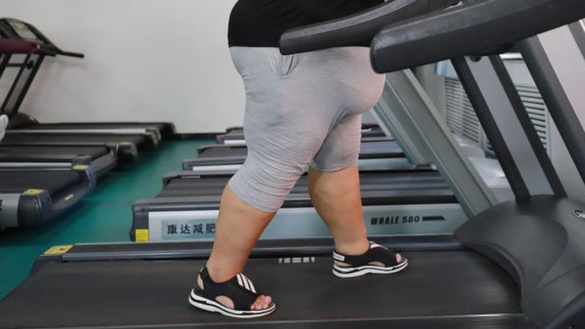 An overweight woman wearing grey shorts and a black t-shirt walks on a treadmill