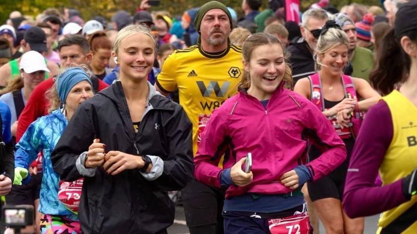 A group of runners and spectators at the start of the race. They are all smiling as they set off. At the front are two young women, one with blonde hair and a black running jacket, the other with brown hair and a dark-pink jacket.