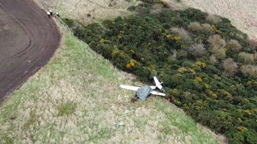 A  bird's eye view of the wrecked white plane in a field. To the top are dark green bushes, dry white grass at the bottom of shot, and the corner of a muddy brown field top left, with three small figures wearing hi vis walking along the edge. 