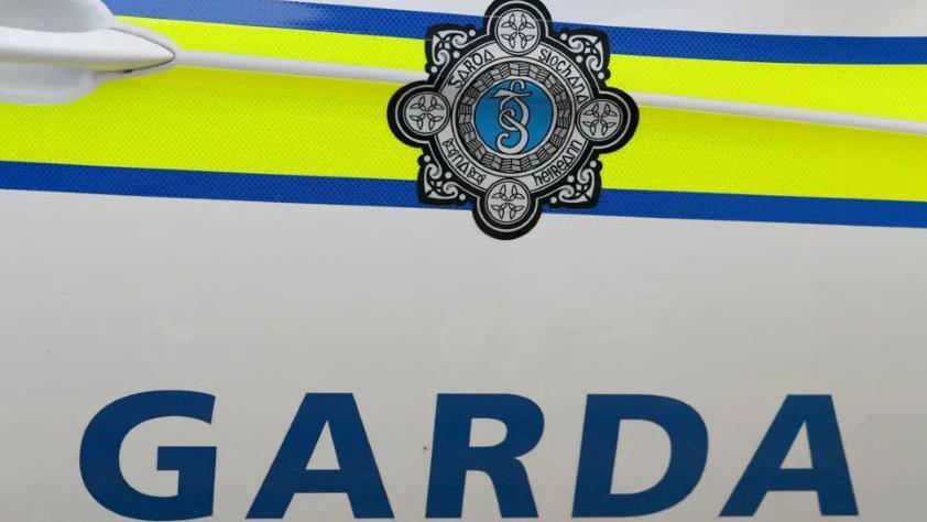 The image depicts the side of a white An Garda Síochána, Irish police force, car with the words garda written in blue capital letters. Above the word are blue and yellow lines with the crest of the organisation also visible in the middle of the lines.