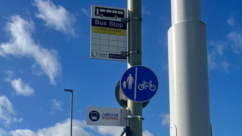 A bright blue sky in the background. In the foreground there's a zoomed in view of a lampost and a pole with a bus stop sign and National Express sign. There is also a sign on the pole showing where pedestrians and cyclists should position themselves while using the pavement.