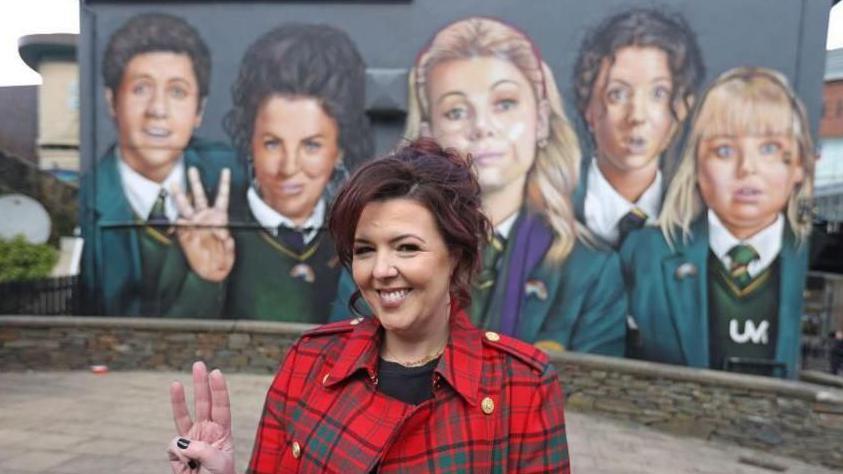 Derry Girls creator Lisa McGee, in red and green check jacket and holding up three fingers on her right hand, stands in front of the Derry Girls mural. The mural depicts the characters from her hit comedy show.
