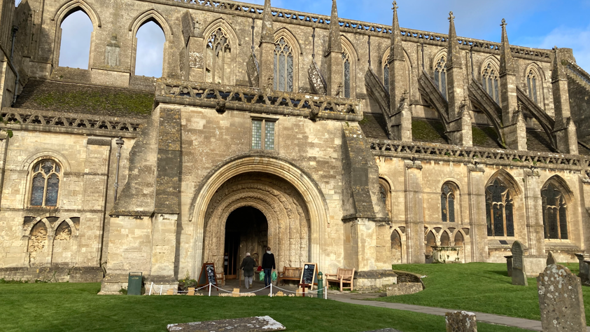 An exterior view of Malmesbury Abbey showing the entrance way, the arched windows and the buttresses. The church yard and several gravestones are also visible.   