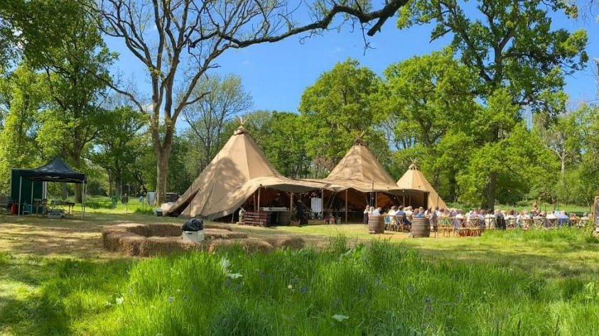 Tipis in the woodland at the estate, with people sat at tables outside them 