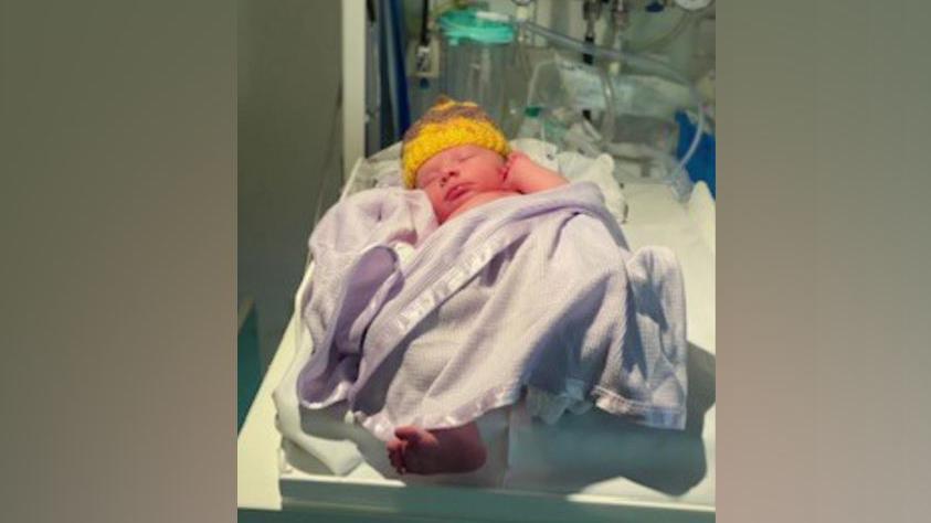 Teddy as a baby in hospital. He is wearing a yellow hat and shrouded in a white blanket.