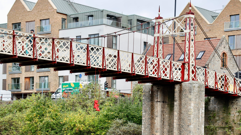 A sculpture of a bright red teddy bear on a red swing can be seen hanging from the bridge. The red and white bridge can be seen in the picture, as well as buildings and the road behind it. 