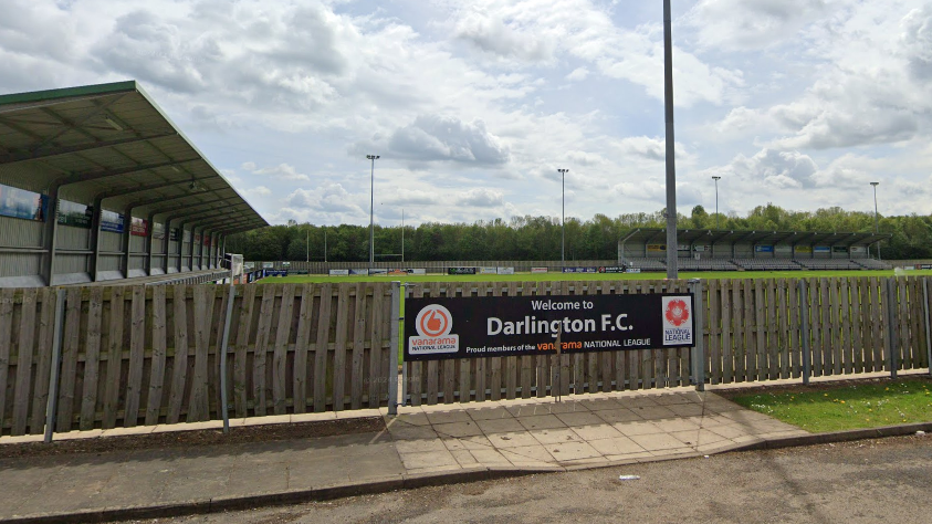 Blackwell Meadows is a football pitch with a wooden fence surrounding it. A sign that reads 'Welcome to Darlington FC' with various sponsorships can be seen on the fence in front. Spectator stands are in the distance.