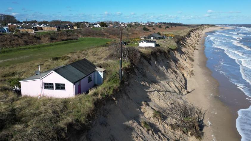 A pink chalet on the edge of a sandy cliff that drops down to the beach. Other chalets can be seen in the background.