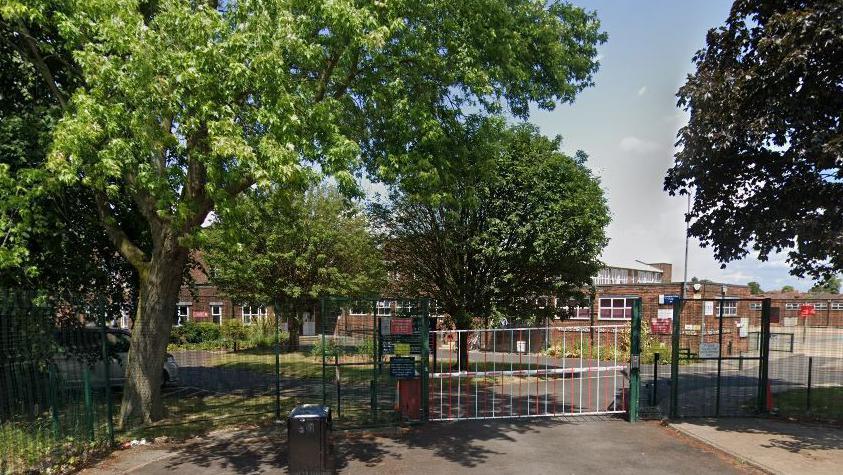 The gated entrance to the school with trees on either side. The red brick building can be seen stretching across the back.