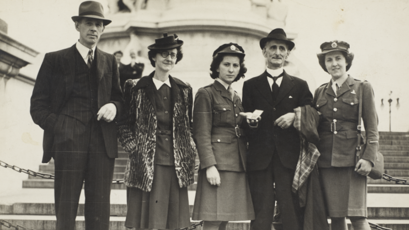 Black and white photo of five people dressed smartly in front of London's Trafalgar Square. On the left is a man in a suit and a hat. In the middle are two women, the second one wearing a military uniform. Next to her is another man in a black suit holding the Victoria Cross medal. On the far right is a woman in a military uniform. 