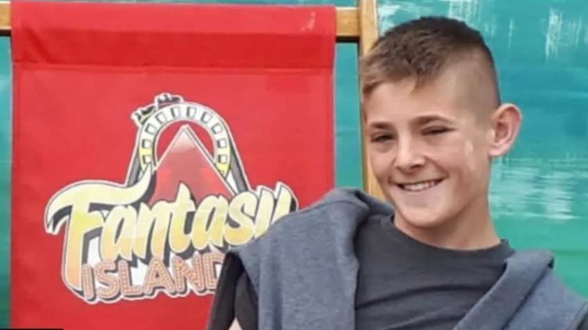 A picture of a young boy smiling wearing a grey t-shirt with a grey jumper resting on his shoulders. He is standing in front of a red sign which reads 'Fantasy Island'