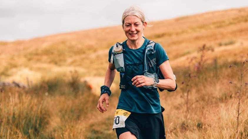 A woman runs along a narrow trail through a dry, grassy field, wearing a blue shirt, black shorts, and trail running shoes. She carries a hydration vest with visible water bottles and a race bib pinned to their shorts.