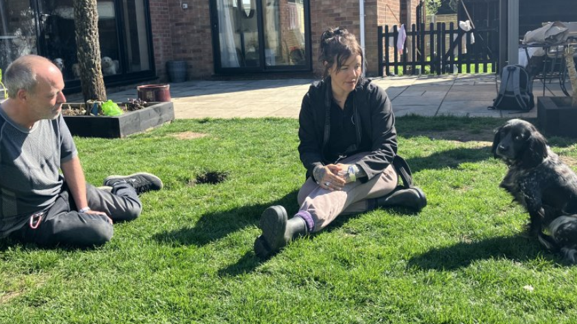 A man with grey hair, wearing a grey T-shirt and jeans, sits on a green lawn next to a woman with brown hair wearing a black jacket, sand-coloured slacks and wellies. To the right, a black cocker spaniel looks towards the camera.