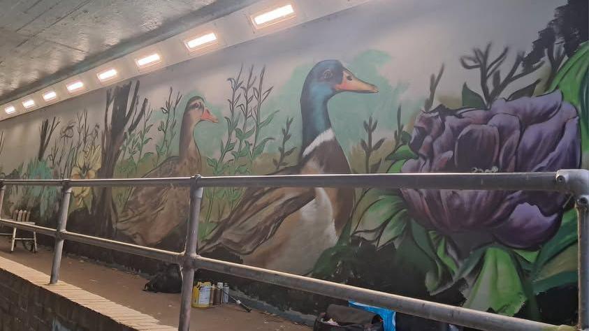 Duck, purple flowers and green plants painted on the side wall of an underpass. There is a small wall with metal railings on top in front of it.