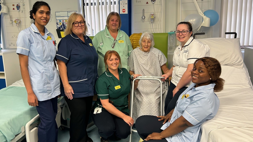 Gloria Malpass-Leek is sitting on a chair on a ward with a frame in front of her and six nursing staff in uniform sitting around her.