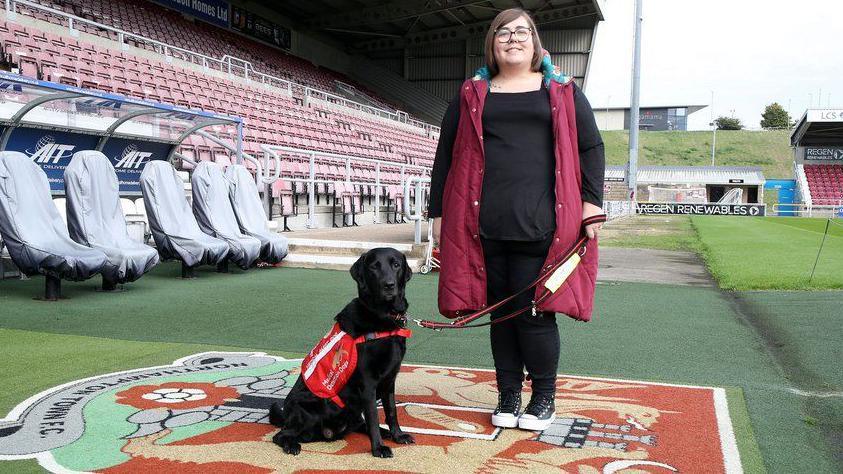 A woman with a black top and black jeans and a maroon long gilet stands next to a black Labrador dog in front of the dugout at Sixfields stadium. 