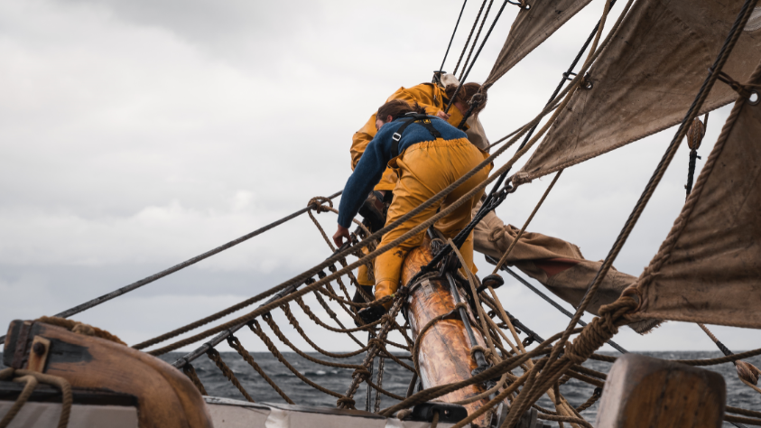 Crew members in yellow wet weather gear work the rigging on the bow of a traditional tall ship. 