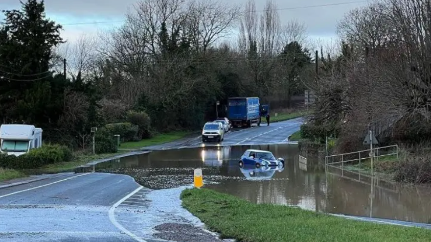 A flooded road with a blue mini abandoned in the waters and several other vehicles parked nearby, with one about the enter the floods