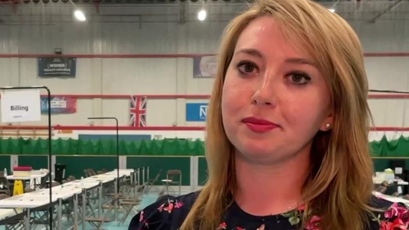Sarah Bool has fair shoulder-length hair and is wearing a black, red and green floral dress. She has a pensive expression and is standing in front of empty tables and chairs at the election cunt in some kind of sports hall.