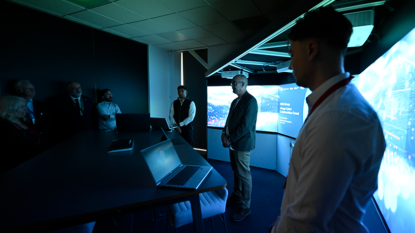 A group of people dressed in suits in one of the cyber security labs at Herdus House. The room is dark and has large screens all across one wall and laptops on a table in the middle of the room.