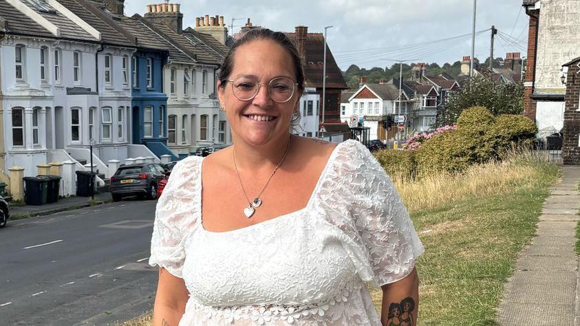 Kelly Warwick standing outside her house in Brighton. She wears a white blouse and dark jeans. She has dark hair tied up, and glasses.