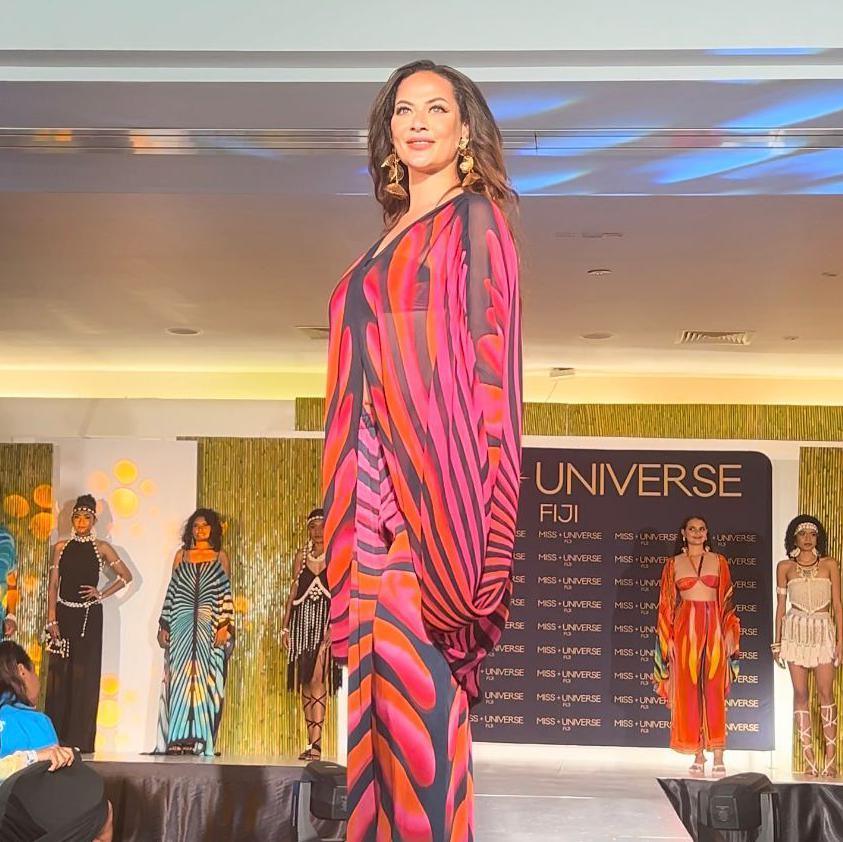 A woman in the foreground wearing a red, pink, black and orange outfit poses on stage. Behind her in the background are her seven fellow contestants for the Miss Fiji contest.