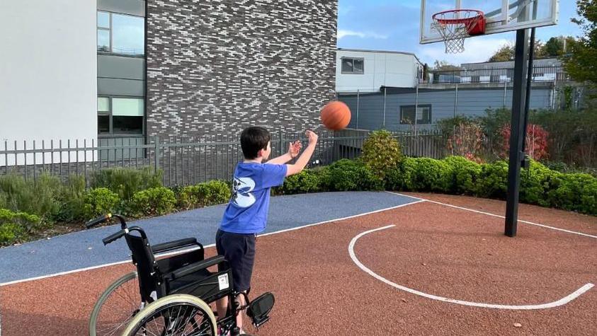 Harry is seen in a basketball court standing up from his wheelchair in an attempt to shoot the ball through the hoop.