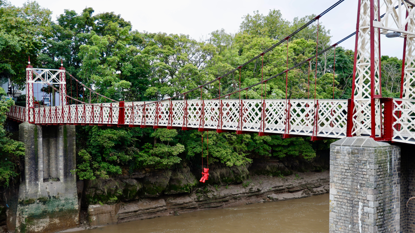 A sculpture of a bright red teddy bear on a red swing can be seen hanging from the bridge above the water. The bridge is a red and white footbridge, and is seen in front of the riverbank and overhanging trees. 