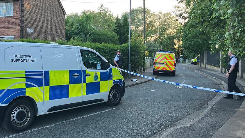 Merseyside Police officers and vehicles inside a cordon following an incident around the Tobruck Road area in Huyton on a sunny evening.