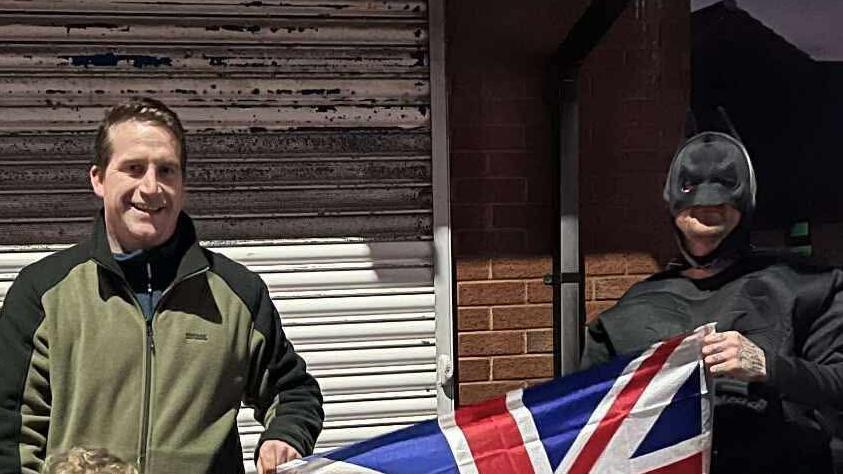 Councillor Steve Jones and a man in Batman suit smile at the camera, holding a Union flag between them