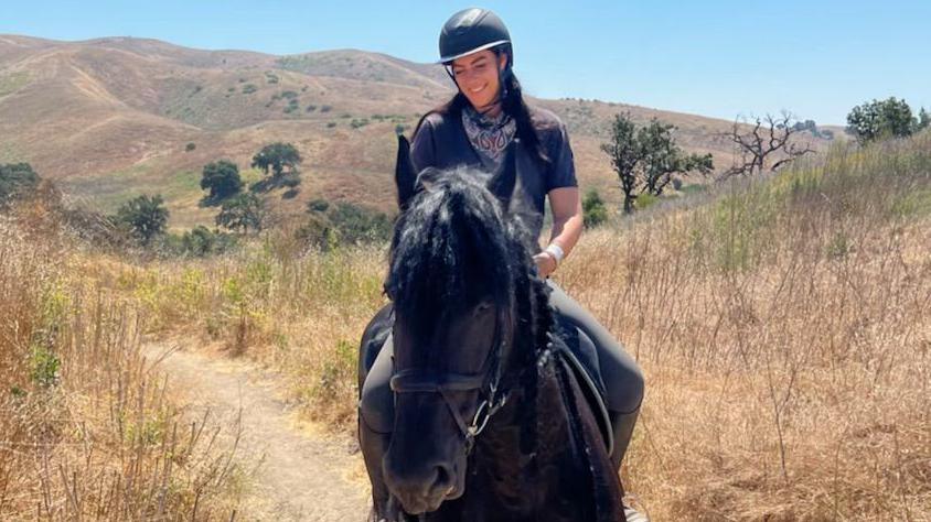 Thea Creasy rides a black horse through a field. She wears a black riding helmet, T-shirt and neckerchief. The horse walks on a path. The fields surrounding them have some greenery but are largely brown from the sun.