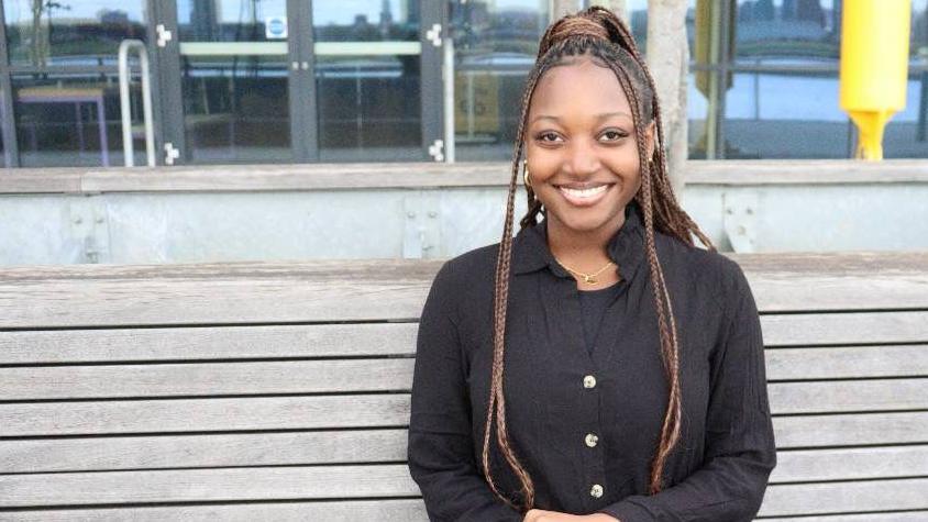 A black woman with braids, wearing a black shirt and trousers, smiles towards the camera as she sits on a wooden bench