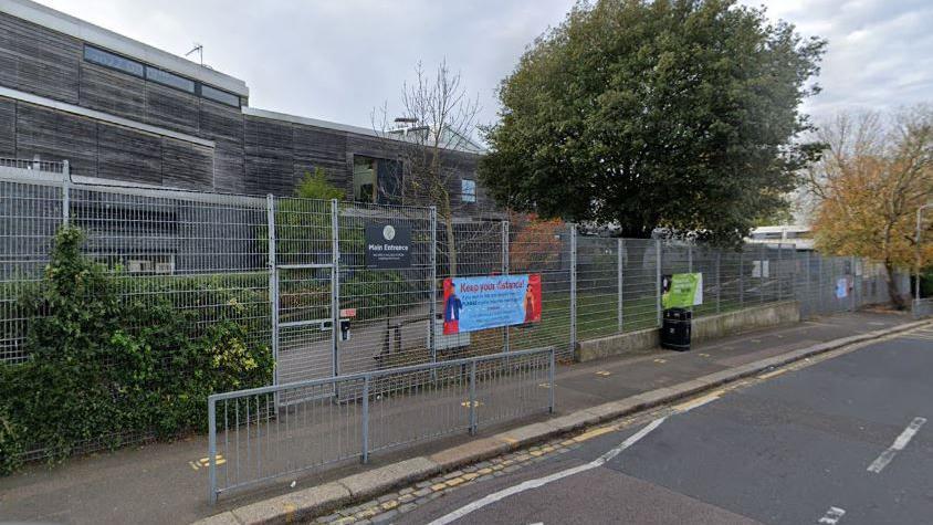 StreetView image showing the main entrance to Mulgrave Primary School, a building clad in wood with high metal fencing and gates surrounding the boundary