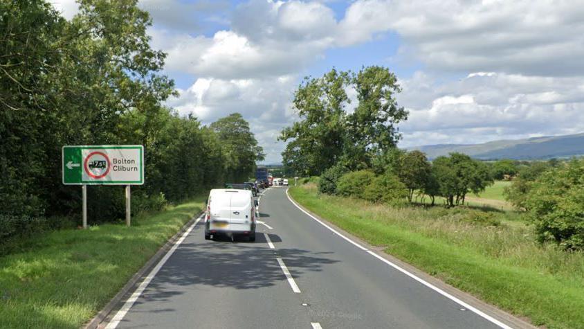 Queuing traffic on a section of road, with green, leafy verges. There is a motoring sign with a left-pointing arrow, saying Bolton and Cliburn.