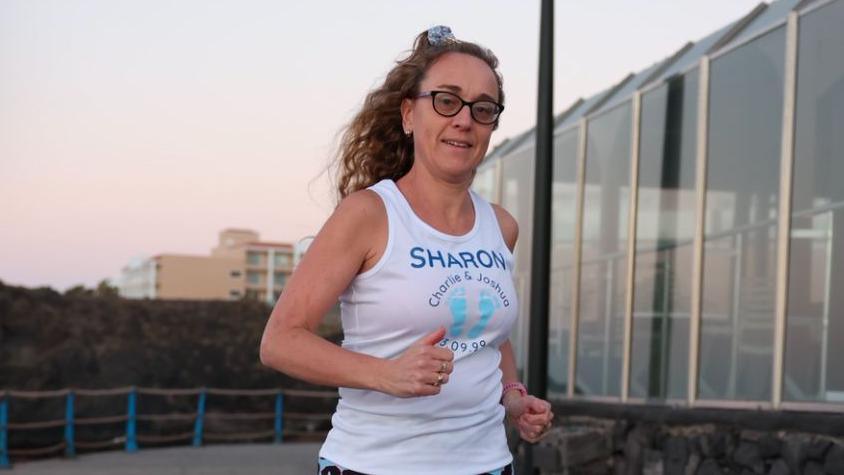 Sharon Darke with long hair wearing a white T-shirt with her first name on the front along with the names of her twin boys. She is running past a building with several full-length windows. A block of flats is in the distance behind her