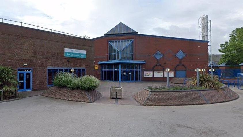 An image of Goole Leisure Centre taken from Google StreetView. It is a red brick building with bright blue windows and doors. There are raised beds with shrubs in, built in the same bricks