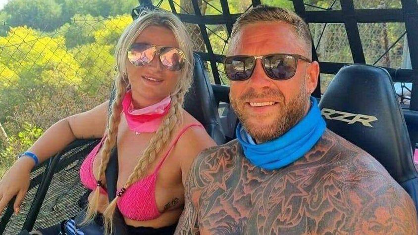 Aaron and Samantha Morris on holiday. He has his top off showing a body full of tattoos. Both are smiling and wearing sunglasses. Samantha has blonde hair.