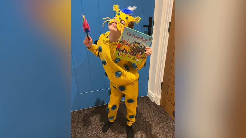 A child dressed as The Tickle Monster in a bright yellow costume with green pompoms on and a yellow mask. The child is also holding a book called Tickle Book. 