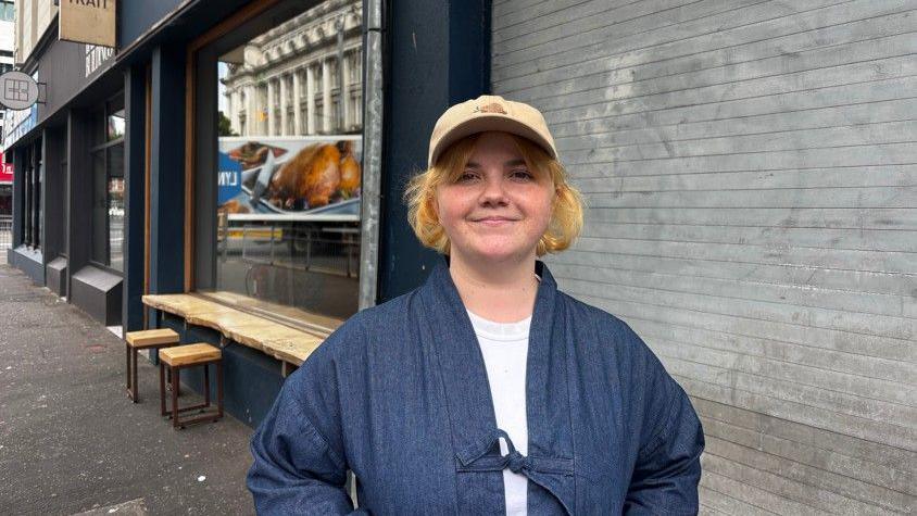 A young girl on the street, smiling. Wearing a white top, denim jacket and yellow cap.